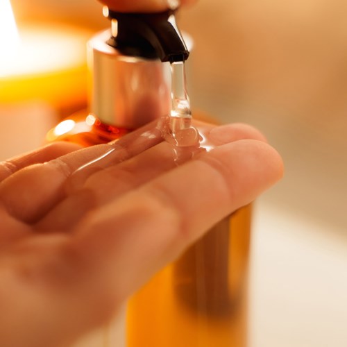 A hand is under a silver soap dispenser, with soap falling onto the palm. Warm lighting creates a cozy feel.