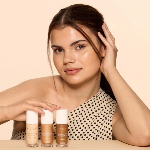 A woman with long brown hair and light skin touches her face, next to three bottles of skincare products.