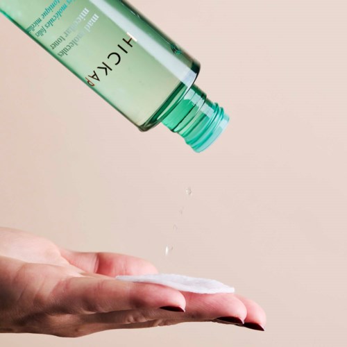 A person using a cotton pad with a green HICARE bottle, likely skincare or cleaning product. No background details.