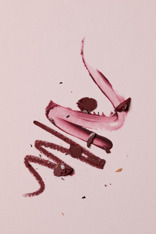 A lipstick stain with smudges and streaks on pink paper, resembling a face with a closed eye and lips.