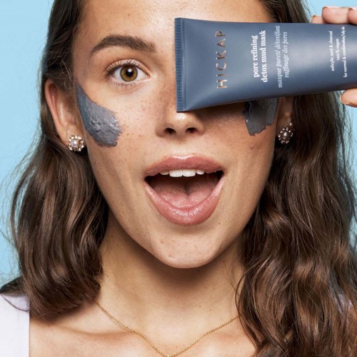 A woman with brown hair and earrings, showing surprise, holds a blue tube near her face, with grey face mask on her cheek.