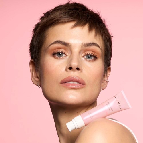 Een vrouw met kort haar en make-up houdt een roze tube tegen een roze achtergrond.