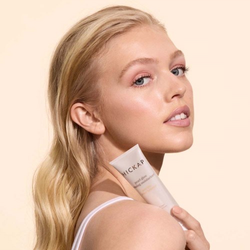 A woman with long blonde hair holds a tube labeled 'HICKAP'. She looks over her shoulder with clear skin and a natural look.