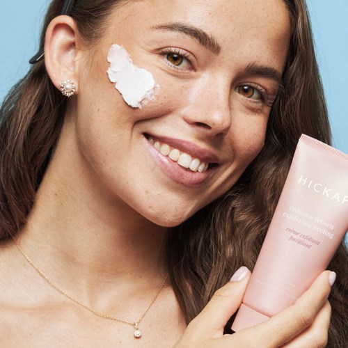 A woman with a smile applying cream on her cheek, holding a skincare product, with clear skin and earrings.