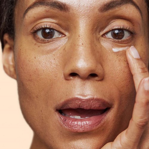 A woman applies eye cream with her finger, close-up of her face showing clear skin and brown eyes.