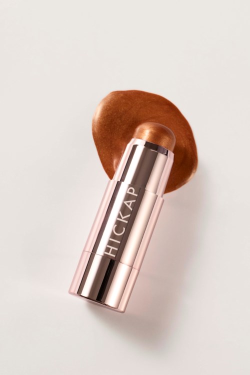 A makeup stick with a label that says HICKAP, placed on a brown cushion. The stick is rose gold-colored.