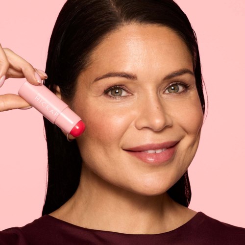 Frau lächelt, hält eine pinke Kapsel vor ihr Gesicht, vor rosa Hintergrund.