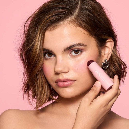Eine Frau mit schulterlangem, welligem Haar verwendet ein rosa Beauty-Tool auf ihrer Wange vor pinkem Hintergrund.