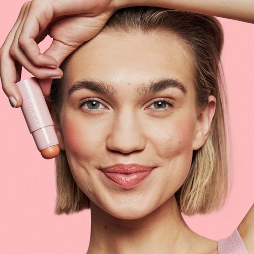 Eine junge Frau mit blonden Haaren und klarer Haut lächelt, während sie ein pinkes Hautpflegeprodukt in der Nähe ihres Gesichts hält.