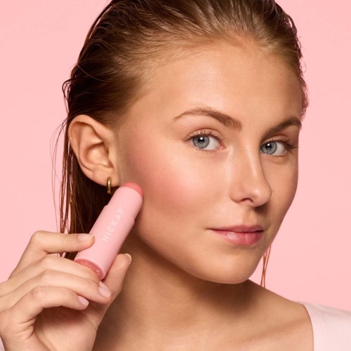 Eine Frau benutzt ein rosa Hautpflegegerät an ihrer Wange, mit neutralem Ausdruck und rosa Hintergrund.