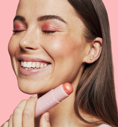Smiling woman with closed eyes applies lipstick on her lips, holding a pink lipstick tube against a pink background.