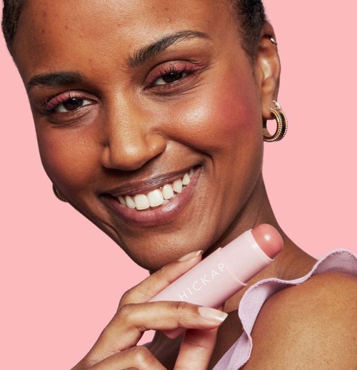 Eine Frau mit dunkler Haut und Ohrringen hat rosa Hintergrund und hält einen rosa Lippenstift.