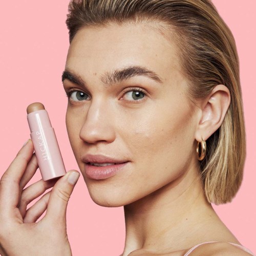 A woman holds a lip balm tube close to her face, smiling slightly. She has blonde hair, blue eyes, and wears a pink top.