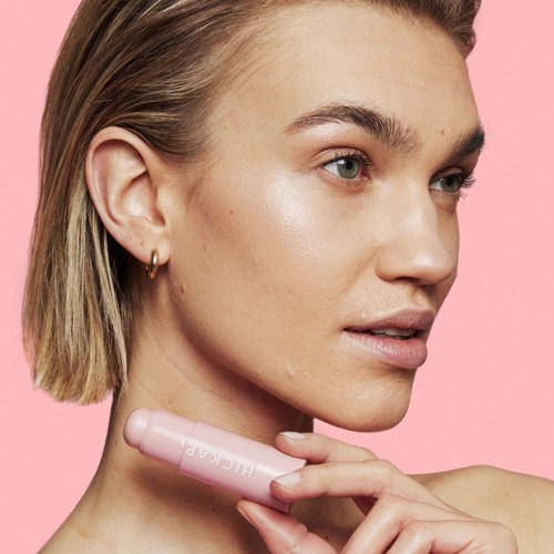 Eine Frau mit kurzen blonden Haaren und heller Haut trägt ein Hautpflege- oder Make-up-Produkt am Hals auf vor rosa Hintergrund.