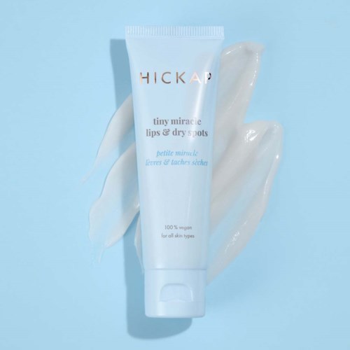 A white tube of Hicksap tiny miracle lips and dry spots sits on a light blue background with some cream swipes.