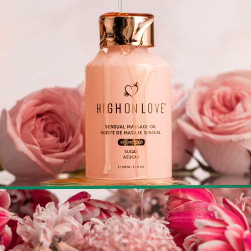 Eine rosa Flasche mit Massageöl und goldener Kappe, umgeben von rosa Rosen und Blumen. Das Etikett zeigt Marke und Produktdetails.