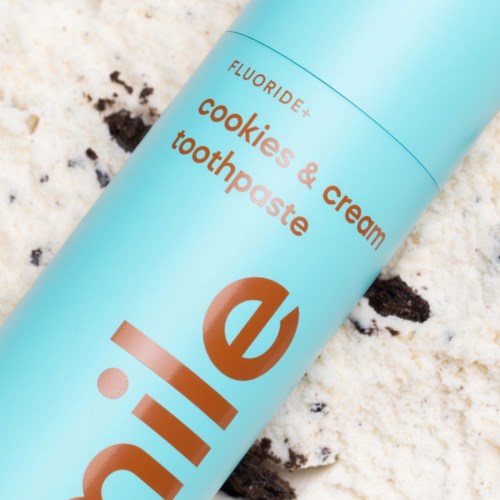 Lichtblauwe tube met label "Cookies & Cream" tandpasta, met wat koekjes op het oppervlak in de buurt.