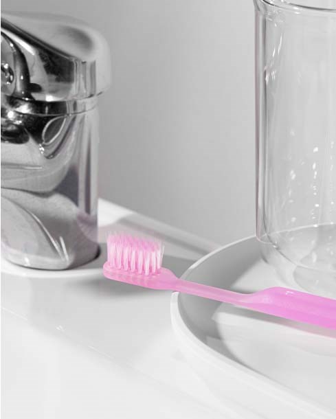 A pink toothbrush rests on a white plate next to a glass jar, with a water tap visible in the background.