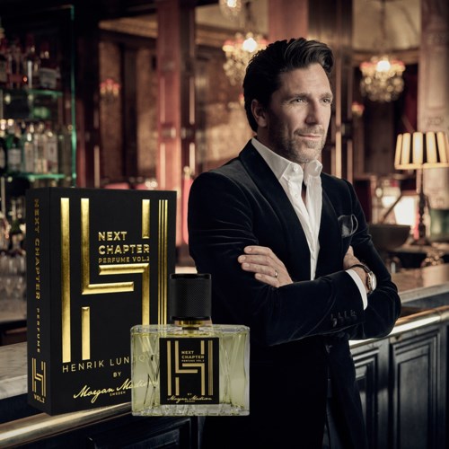 En man i en svart sammetkostym står med armarna i kors bakom en parfymutställning i ett elegant, varmt belyst rum.