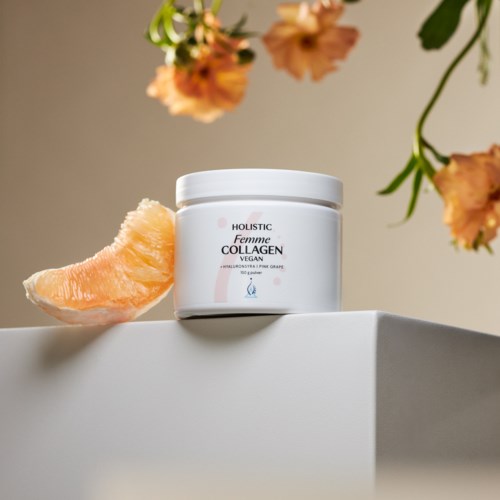 En vit burk märkt 'Femme Collagen Vegan' på en vit yta, med en rosa frukt och orange blommor i närheten.