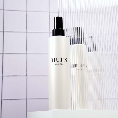 A white bottle labeled 'HUFS ALL IN ONE' is on a white surface, with some tall, thin bottles beside it.