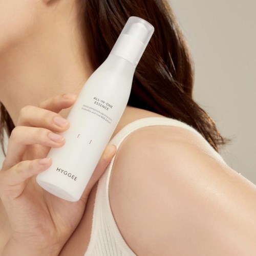 A woman holds a white skincare bottle near her shoulder, showing healthy, smooth skin. The background is neutral.