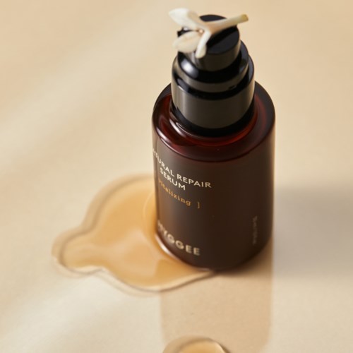 A brown bottle with a pump, labeled 'Natural Repair Serum,' sits on a cream surface with spilled product nearby.