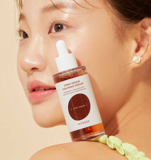A woman holds a skincare serum bottle near her face, with clear skin and minimal makeup, smiling slightly.