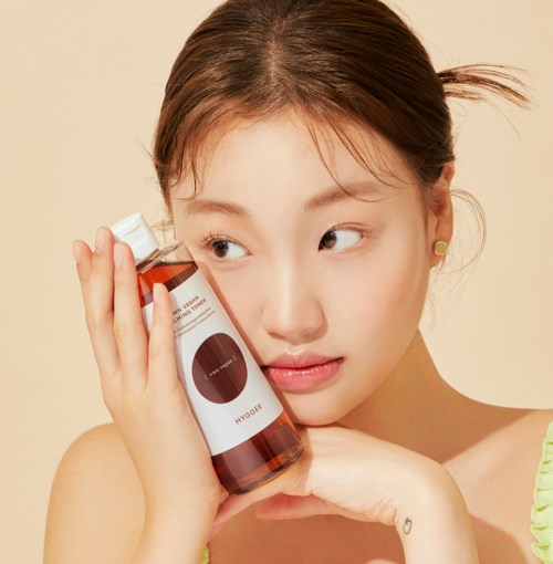 A young woman holds a bottle of skincare product close to her face, looking thoughtful and slightly smiling.