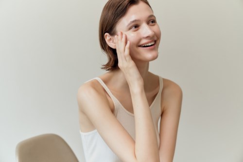 A smiling woman with short brown hair sits at a table, touching her face with hand, in a light-colored tank top.