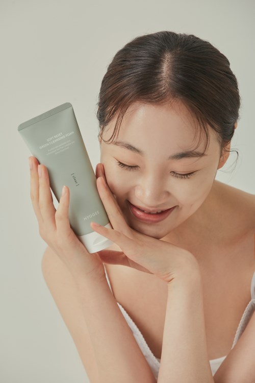 A woman happily holds a gray skincare tube near her face, smiling softly with her eyes closed.
