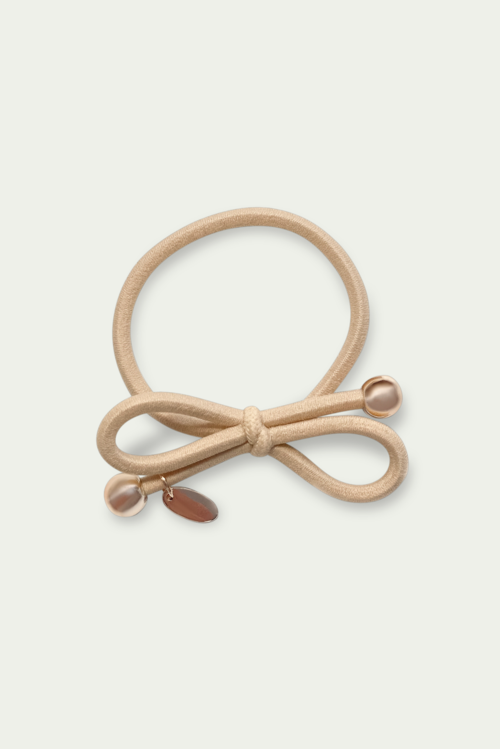 A beige hair tie with a bow and small decorative charms, including a pearl and a gold-colored charm.