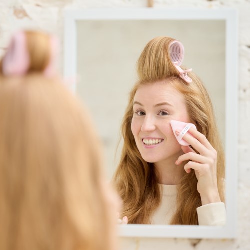 A woman with red hair uses a pink hair roller and a sponge to clean her face, smiling at the mirror.