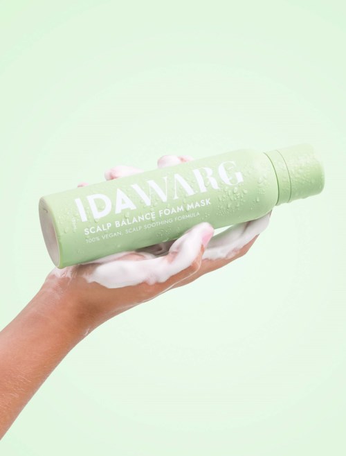 En hand håller en grön flaska med IDAARG scalp foam mask mot en ljusgrön bakgrund.