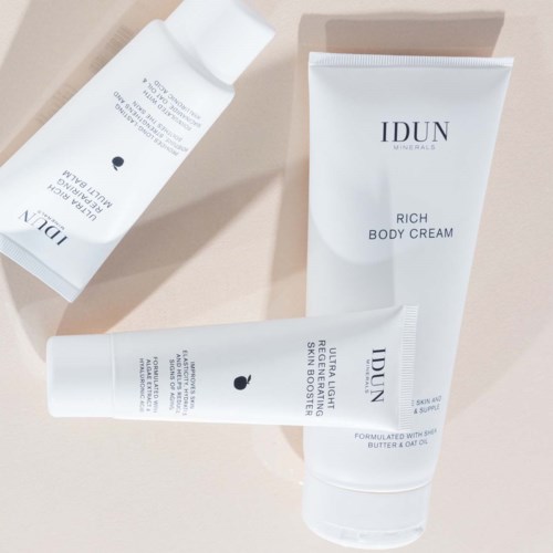 Three white tubes of skincare products with black text, set on a beige surface. The products are from IDUN. There is a body cream, a face serum, and a lightweight serum.