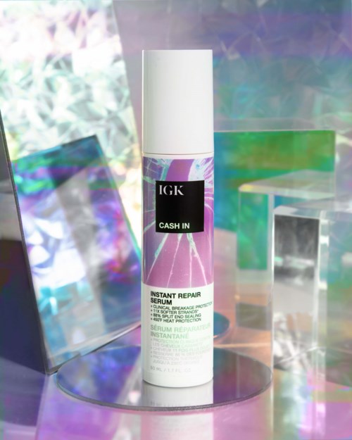 Een witte fles met het label IGK Cash In, een direct reparatieserum, staat tussen kleurrijke, spiegelende oppervlakken of panelen.