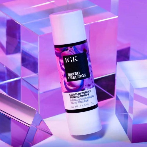 Eine weiße Flasche IGK Mixed Feelings Leave-in-Pflege, lila Toning Drops, vor einem reflektierenden lila und pinken Hintergrund.