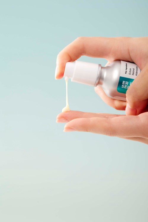 Een hand houdt een klein flesje dat crème of gel afgeeft op de andere hand. Lichtende achtergrond met zachte kleuren.