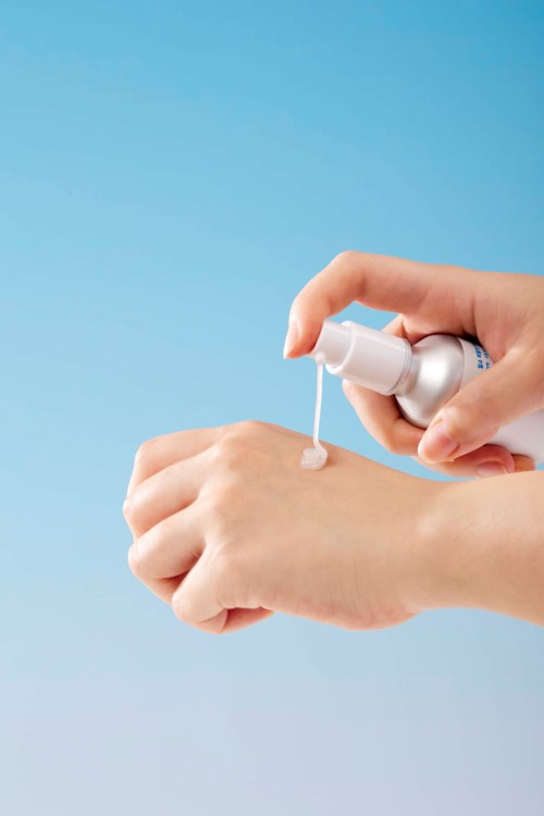 Een persoon brengt een roze-achtige crème of lotion aan op de hand met een sprayfles.
