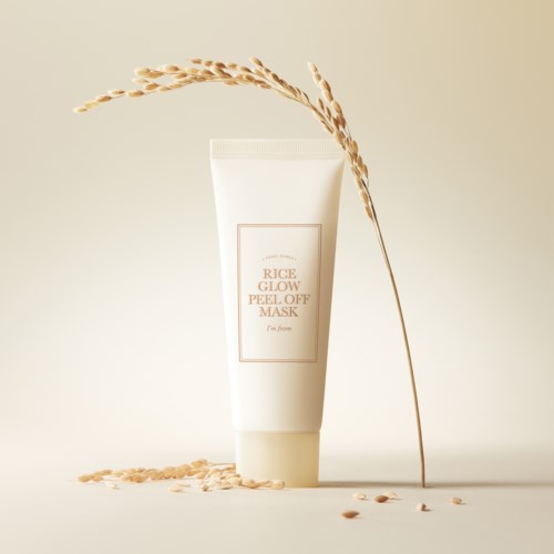Een witte tube met Rice Glow Peel-Off Mask, omgeven door rijstkorrels en een droog plantenstengel die erop leunt.