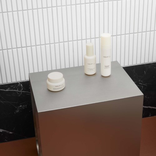 Three white cosmetic bottles and a jar are placed on a gray display stand against a white tiled wall.