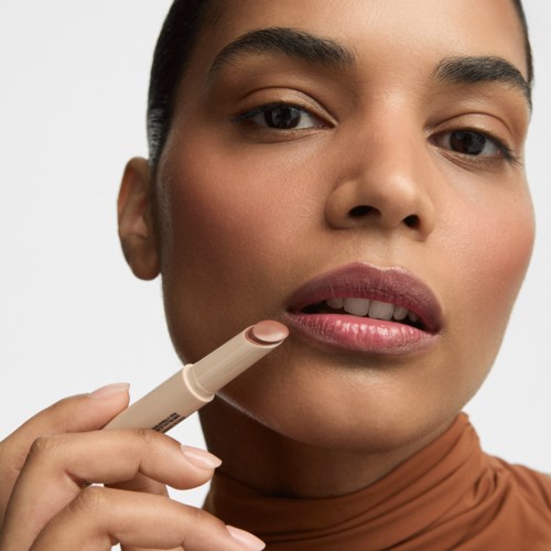 A woman applying lipstick, close-up of her lips and face with neutral makeup, in front of a plain background.