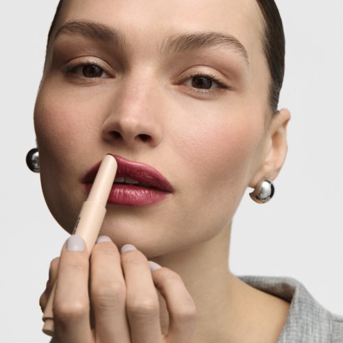 Close-up of a woman applying lipstick with a beige lipstick pencil, wearing earrings and a light gray top.