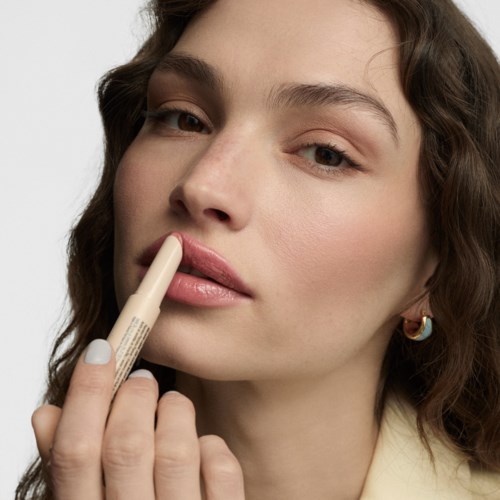 A woman applying light pink lip gloss with her fingers, wearing earrings and a beige top.