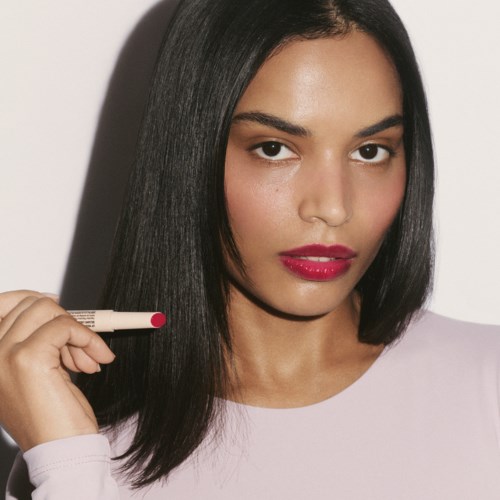 Woman with straight black hair wearing a light pink top, holding a lipstick near her face, with red lips.