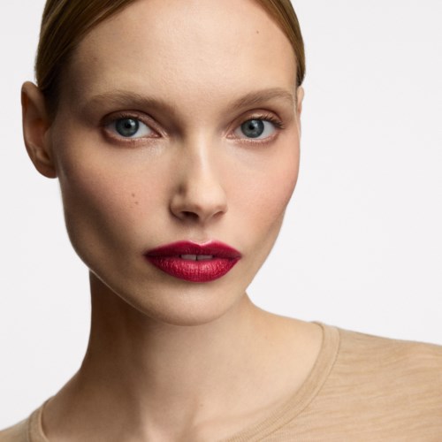 Eine junge Frau mit roten Lippenstift und blauen Augen, schaut direkt in die Kamera, trägt ein beiges Oberteil.