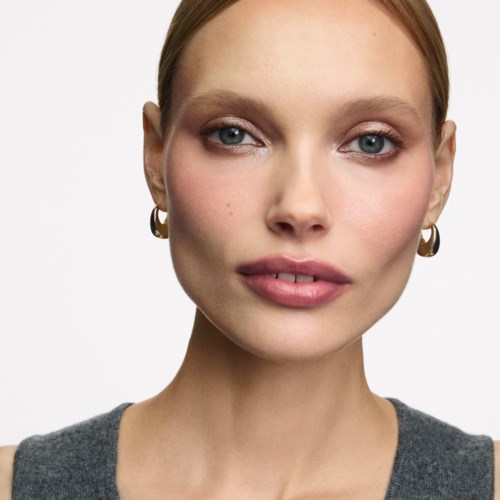 Eine Frau mit heller Haut, rotem Haar, blauen Augen, goldene Ohrringe und neutralem Ausdruck.