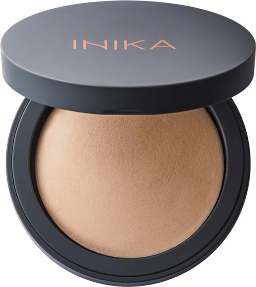 A compact with a beige powder inside and a dark gray lid labeled 'INIKA.'