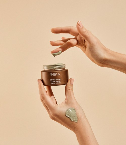 Dłonie trzymają mały słoik, z zieloną maseczką, która jest nakładana. Na słoiku jest napis 'INIKA' i 'Detoxifying Clay Mask.'
