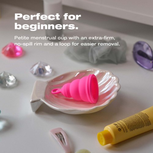 Eine kleine rosa Menstruationskappe auf einem weißen Teller, mit einer gelben Tube und Dekokristallen.